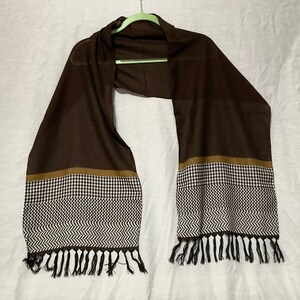 May include: A brown scarf with a black and white geometric pattern and brown trim. The scarf has black tassels on the ends.
