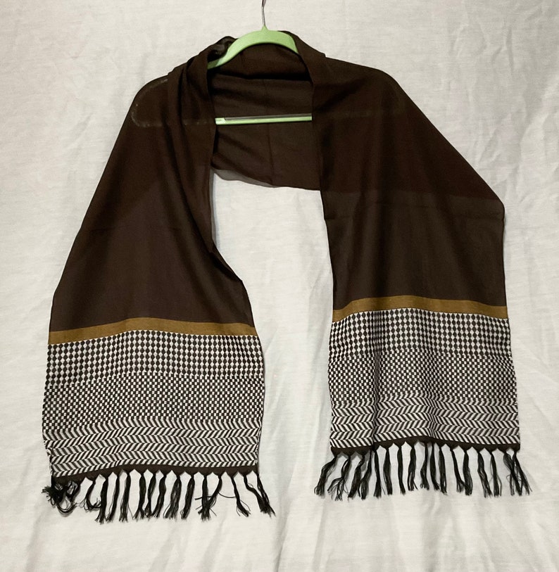 May include: A brown scarf with a black and white geometric pattern and brown trim. The scarf has black tassels on the ends.