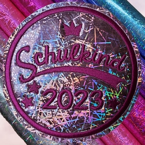 Könnte beinhalten: Ein runder, bestickter Aufnäher mit einem holografischen silbernen Hintergrund im Scherbenmuster. Der Aufnäher hat einen dunkelvioletten gestickten Rand und zeigt den Text Schulkind 2023 in dunkelviolettem Garn, verziert mit einer kleinen Krone und Sternen.