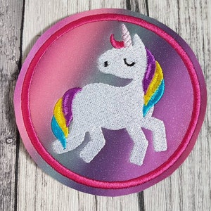 Könnte beinhalten: Ein weißes Einhorn mit einer Regenbogenmähne und einem Regenbogen-Schwanz ist auf einem rosa und violetten Kreis mit rosa Rand gestickt.