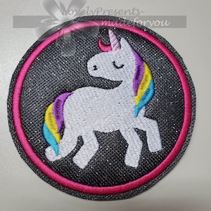 Könnte beinhalten: Ein runder Patch aus schwarzem Glitzerstoff mit pinkfarbenem Rand, auf dem ein weißes Einhorn mit Regenbogenmähne und -schwanz gestickt ist.