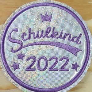 Könnte beinhalten: Ein weißer, runder, glitzernder Patch mit einem lila gestickten Rand. Der Patch zeigt eine lila Krone, den Text "Schulkind 2022" und zwei lila Sterne.