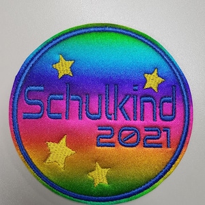 Könnte beinhalten: Ein kreisförmiger Aufnäher mit einem regenbogenfarbenen Hintergrund. Der Text "Schulkind" und "2021" ist in blauem Garn gestickt. Vier gelbe Sterne sind um den Text herum angeordnet, alles eingefasst von einem blauen Rand.