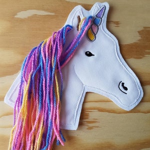 Könnte beinhalten: Weißer Filz-Einhornkopf mit einer Regenbogen-Garnmähne. Das Einhorn hat ein Horn mit einem lila, blauen und gelben Design.