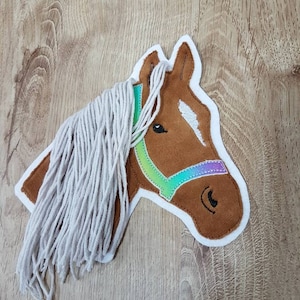 Könnte beinhalten: Ein brauner Pferdekopf mit einem weißen Streifen im Gesicht, einem regenbogenfarbenen Zaumzeug und einer langen weißen Mähne. Der Pferdekopf ist aus Filz und ein Dekorationsartikel.