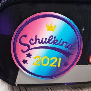 Könnte beinhalten: Ein kreisförmiger, bestickter Aufnäher auf einem dunklen Rucksack mit einem Farbverlauf in Pink, Lila und Blau. Der Aufnäher zeigt das Wort Schulkind in violetter Schreibschrift, eine gelbe Krone, die Jahreszahl 2021 in Gelb und kleine lila Sterne.