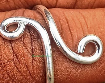 May include: A silver-colored ring with a unique, swirling design. The ring features two curved lines that meet, with a looped end on each side. The ring is worn on a finger, showcasing its intricate, minimalist style.