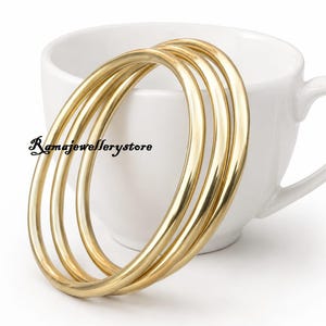 May include: Three polished gold-tone metal bangles with a smooth, rounded surface. The bracelets are stacked together, displaying a reflective finish. The text "Ramajewellerystore" is printed in black on the left side of the bangles.