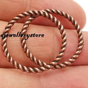 May include: Two copper-colored, twisted metal rings are displayed. The rings are circular and have a rope-like texture. The rings are held in a person's hand. The text "Ramajewellerystore" is visible in the image.