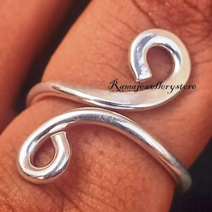 Può includere: Un anello color argento con un design a spirale. L'anello presenta due estremità arrotondate a forma di spirale collegate da una fascia curva. L'anello è su un dito, con il testo "Ramajewellerystore" visibile.
