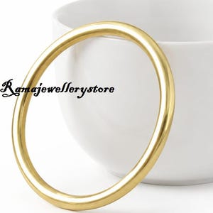 May include: A smooth, polished gold bangle bracelet with a rounded profile. The bangle is positioned in front of a plain white ceramic coffee mug. The text Ramajewellerystore is printed in black script on the left side of the gold bangle.