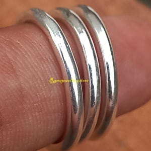 Puede incluir: Primer plano de cuatro anillos de plata apilados en un dedo. Los anillos son lisos y brillantes, reflejando la luz. Los anillos son bandas simples, probablemente de plata de ley. El fondo es una superficie borrosa de tonos cálidos.