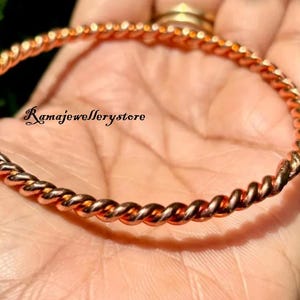Puede incluir: Una pulsera de brazalete retorcida de color cobre. La pulsera tiene un diseño de cuerda y se muestra en una mano. El texto "Ramajewellerystore" es visible en la imagen.
