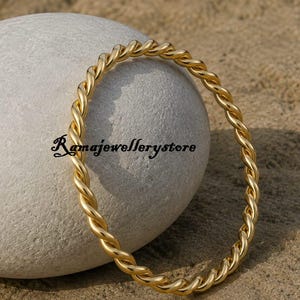 May include: A gold-colored metal bangle bracelet featuring a continuous twisted rope design. The circular jewelry piece has a polished finish and is positioned upright. The text Ramajewellerystore is visible in black script across the center.