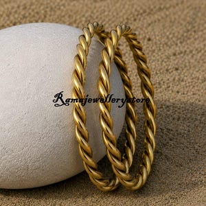 May include: Two gold-toned metal bangles featuring a twisted rope design. The bracelets are positioned against a smooth, light-colored oval stone. The text Ramajewellerystore is visible in black script lettering between the two gold bangles.