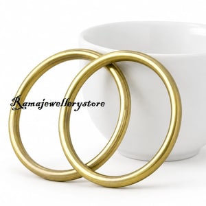 May include: Two smooth, circular gold-toned bangles with a polished finish. The text Ramajewellerystore is printed in black on the left side of the bangles. The bangles are positioned in front of a plain white ceramic cup with a handle.