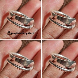 Puede incluir: Un anillo plateado con un diseño único en capas. El anillo presenta tres bandas que se envuelven alrededor del dedo, creando un aspecto moderno y elegante. La imagen es un primer plano que resalta la superficie lisa y pulida del anillo.