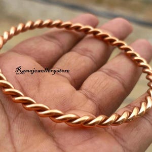 Op de afbeelding: Een koperkleurige armband met een gedraaid touwontwerp. De armband is een doorlopende lus, met een spiraalpatroon. Het oppervlak reflecteert licht, wat de metalen glans benadrukt. De tekst "Ramajewellerystore" is zichtbaar.