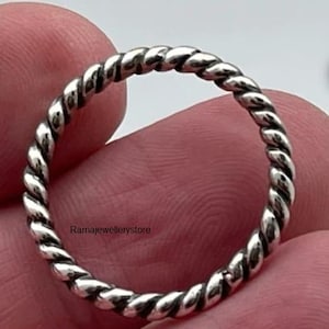 Puede incluir: Un anillo plateado con un diseño de cuerda trenzada. El anillo es una banda simple con un patrón continuo de hebras entrelazadas. El anillo está en la mano de una persona.