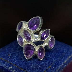 Könnte beinhalten: Ein silberner Ring mit einem Cluster aus Amethyst-Edelsteinen in Tropfenform. Der Ring liegt auf einem blauen Samt-Hintergrund.