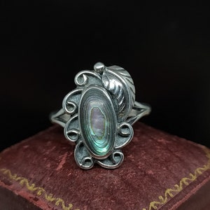 May include: A silver ring with an oval abalone shell inlay and a leaf design. The ring is set on a red velvet box.