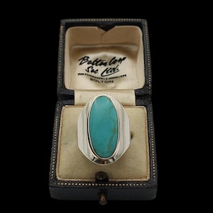 May include: A silver ring featuring a large, oval turquoise gemstone. The ring is presented in a vintage black jewellery box, with the text "Bolton & Son Ltd. Watchmakers & Jewellers Bolton". The ring has a polished silver band.