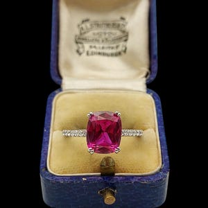 Op de afbeelding: Een zilveren ring met een grote, vierkant geslepen roze edelsteen, gepresenteerd in een vintage blauwe sieradendoos. De ring heeft een bandje bezet met kleine, fonkelende steentjes. De binnenkant van de doos is zachtgeel.