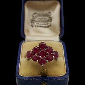 May include: A cluster ring featuring nine round, deep red rubies set in a silver-toned metal frame. The ring is displayed inside an open, rectangular, dark blue velvet jewelry box with a cream-colored satin interior lining and visible text.