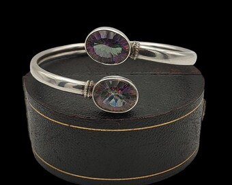 May include: A silver cuff bracelet featuring two oval, iridescent gemstones. The gemstones show a spectrum of colours, including purple, green, and blue. The bracelet is presented on a black box with gold trim.