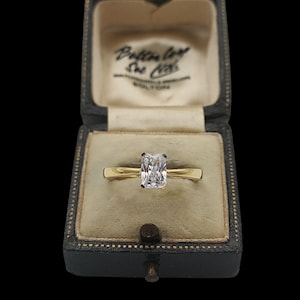 May include: A gold ring with a rectangular, clear gemstone sits in a vintage, dark blue jewellery box. The box has a cream interior and a clasp. The ring's band is thin and simple.