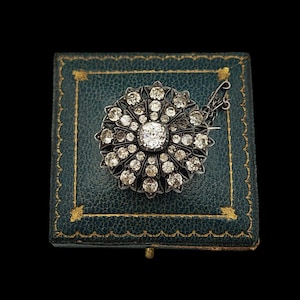 Könnte beinhalten: Vintage silberne Brosche mit einem zentralen, runden Diamanten, umgeben von kleineren Diamanten, in einem floralen Design. Die Brosche liegt auf einer teal-farbenen, antiken Schmuckschatulle mit goldenen Verzierungen, vor schwarzem Hintergrund.