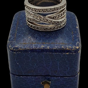 May include: A silver ring with multiple bands, each set with small, clear stones. The bands are interwoven, creating a textured design. The ring sits on a small, blue, leather-like box with a brass clasp, against a black background.