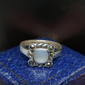 Puede incluir: Un anillo de plata con una piedra blanca en el centro y cuatro piedras negras más pequeñas alrededor. El anillo está sobre una caja de terciopelo azul.