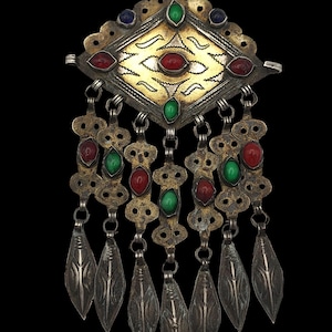May include: A silver pendant with a diamond-shaped top, adorned with red, green, and blue gemstones. The pendant has a gold-colored center and dangling silver leaf-shaped charms, creating an ornate and detailed design. The background is black.