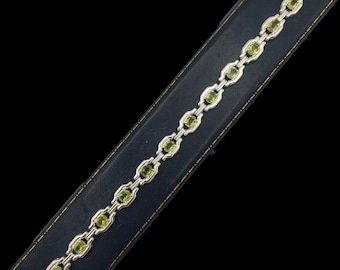 May include: Silver bracelet with oval, light green gemstones. The bracelet's links are interconnected, each holding a gemstone. The clasp is visible. The bracelet is displayed on a dark blue, textured surface. A classic piece of jewellery.