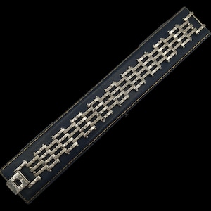 Könnte beinhalten: Ein silberfarbenes Armband mit rechteckigem Verschluss und einer Reihe von miteinander verbundenen rechteckigen Gliedern. Das Armband wird auf einer dunkelblauen rechteckigen Basis mit goldenem Rand vor schwarzem Hintergrund präsentiert.