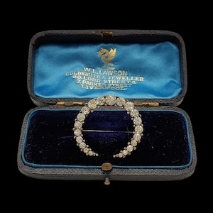 Op de afbeelding: Een vintage, halvemaanvormige broche met talrijke heldere edelstenen, gepresenteerd in een blauw fluwelen doosje. De zwarte doos heeft gouden letters en een logo. Een klassiek sieraad.