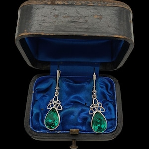 May include: A pair of silver-toned drop earrings featuring faceted emerald green teardrop-shaped stones suspended from intricate silver Celtic knotwork designs. The earrings are displayed inside an open, dark-coloured vintage jewellery box.