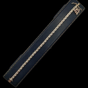 Op de afbeelding: Een gouden armband met talrijke kleine, heldere edelstenen. De armband wordt gepresenteerd in een donkerblauwe rechthoekige doos met gouden randen. Een decoratieve bedel met een hart en vleugels is bevestigd.
