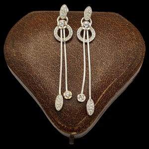 May include: Silver-tone drop earrings with clear, sparkling stones. Each earring has an oval top, a circular design with a central stone, and a chain ending in round and oval stones. The earrings are presented on a brown, textured surface.