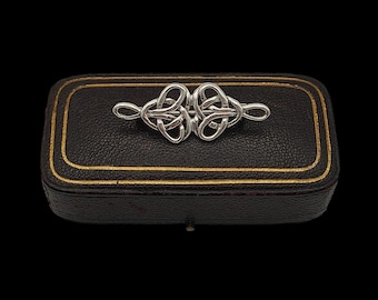 Pode incluir: Um broche prateado com um design de nó celta é exibido em uma pequena caixa preta retangular com bordas douradas. O broche é o ponto focal, mostrando padrões intrincados entrelaçados. A caixa fornece um fundo contrastante.