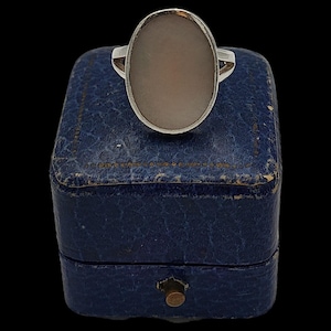 May include: A silver ring featuring an oval, pale stone set in a silver band. The ring is presented on a vintage blue jewellery box with a brass clasp. The background is black.