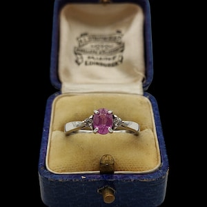 Puede incluir: Un anillo de tono plateado con una piedra preciosa rosa de corte ovalado en el centro, flanqueada por dos pequeñas piedras claras a cada lado. El anillo se presenta en un joyero azul oscuro forrado de terciopelo con el texto A.L. STRUMPF EDINBURGH.