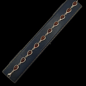May include: A silver bracelet with teardrop-shaped amber stones. The bracelet is presented on a blue, rectangular jewellery box against a black backdrop. The amber stones are a warm, brown colour.