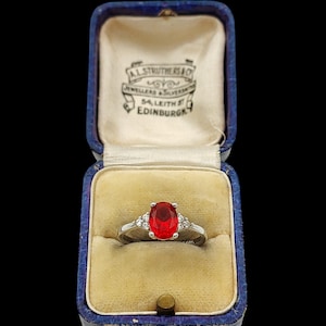 Puede incluir: Un anillo de plata con una piedra preciosa roja central de corte ovalado, flanqueada por tres pequeñas piedras transparentes a cada lado, presentado en un joyero de terciopelo azul con forro interior crema impreso con A.L. STRUTHERS & CO JEWELLERS & SILVERSMITHS 54, LEITH ST EDINBURGH.