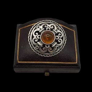 May include: A silver-toned brooch with an amber-colored center stone, set against a dark brown jewelry box with gold trim. The brooch features an ornate, swirling design. The box has a small, brass-colored clasp.