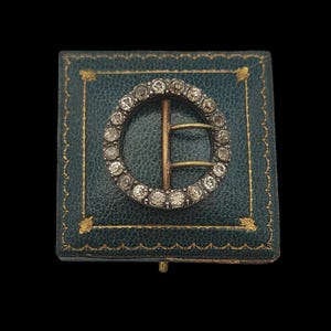 May include: A vintage, circular buckle with a ring of clear stones, set in a silver-toned metal. The buckle has a gold-toned bar and is displayed on a teal, leather-like box with gold trim. The box is closed.
