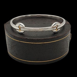 May include: A silver-tone bracelet with gold-colored accents. The bracelet has a hinged design with a clasp. The bracelet is displayed on a dark brown box with gold trim. The background is black.