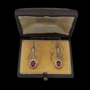 May include: A pair of drop earrings featuring oval-cut deep red gemstones surrounded by small clear stones, set in silver-toned metal. The earrings are displayed inside a dark brown rectangular hinged jewellery box with a beige velvet interior.
