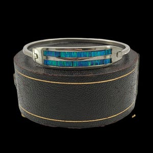 Puede incluir: Una pulsera de plata con una sección rectangular con incrustaciones de vibrantes piedras de ópalo azul y verde. La pulsera se encuentra sobre un fondo oscuro, resaltando su metal pulido y sus coloridos acentos.